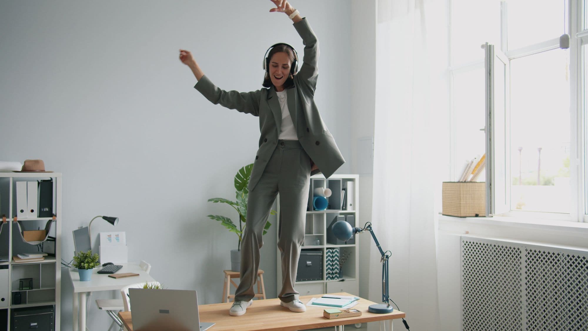 A woman wearing headphones dances exuberantly on her office desk.